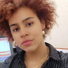 A picture of a tan-skinned woman with a short dark red afro. She is wearing gold hoop earrings and a black collared shirt.
